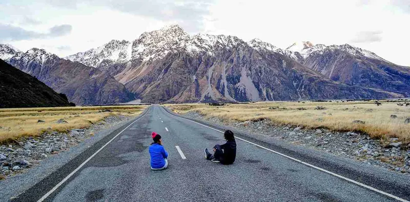 Carretera recta hacia el Mount Cook, con dos personas sentadas en el asfalto