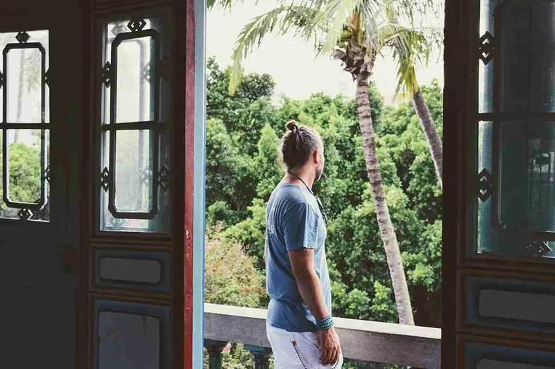 Hombre asomado a una ventana de madera con una palmera al fondo